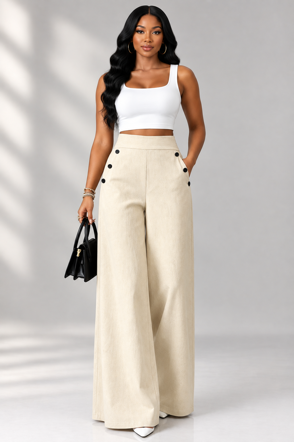 A woman modeling a two-piece outfit consisting of a white crop top and high-waisted, wide-leg beige pants with decorative black buttons on the hips, holding a small black handbag against a studio backdrop with soft window shadows.