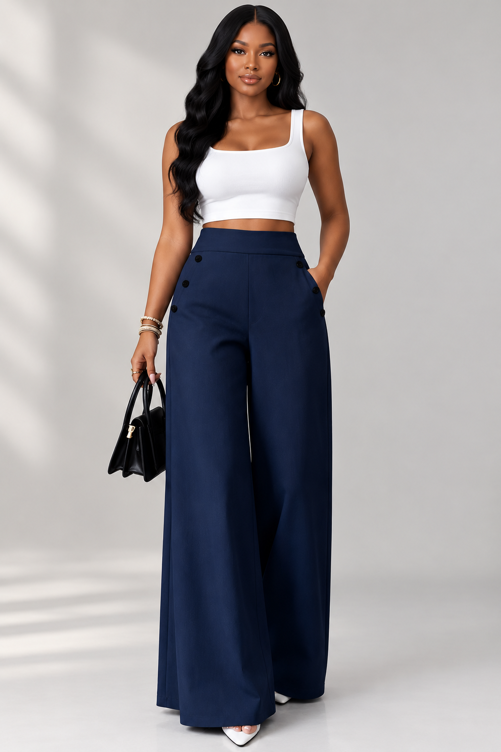 A woman modeling a pair of high-waisted, wide-leg navy blue pants with decorative buttons on the side, paired with a white crop top and holding a black handbag against a light gray studio background.