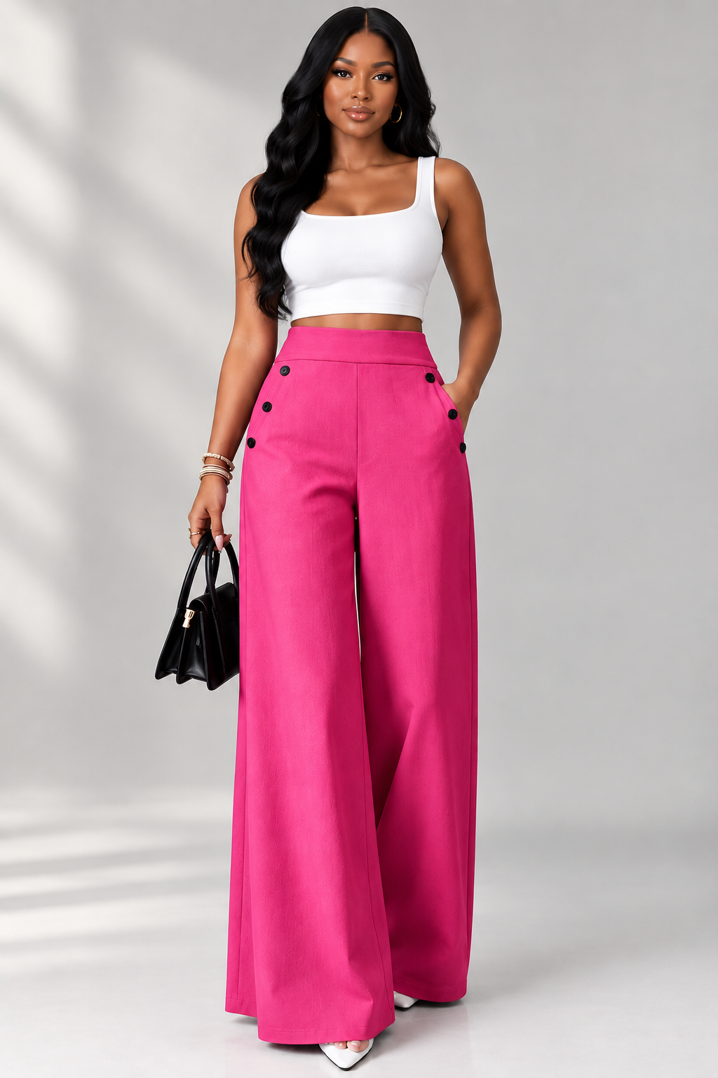 A woman modeling bright pink wide-leg pants with black buttons, paired with a white crop top and holding a black handbag, set against a light gray studio background with soft shadows.
