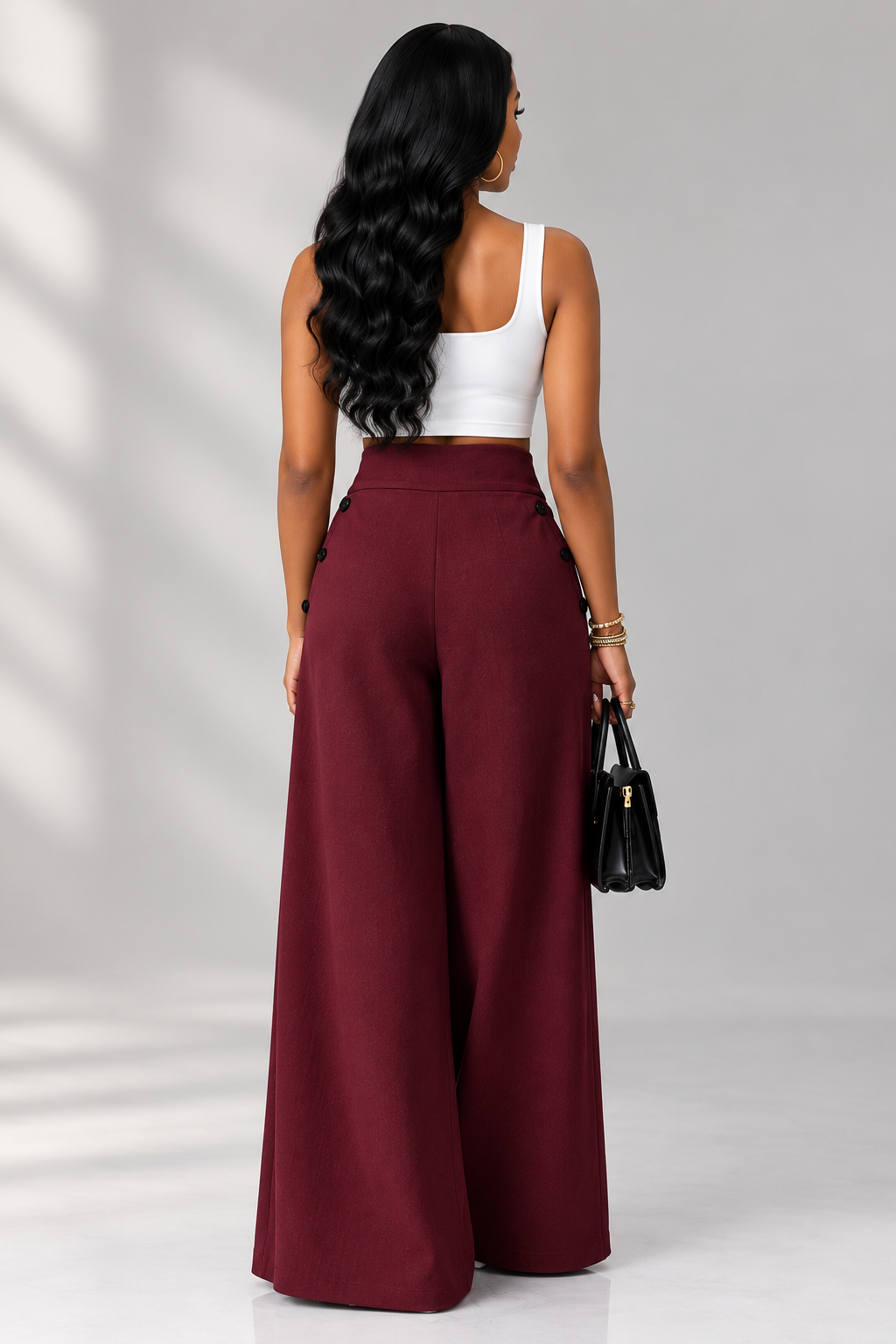 A woman shown from the back wearing a white cropped tank top and high-waisted, wide-leg burgundy pants, holding a small black handbag against a light gray studio background with soft shadow patterns.